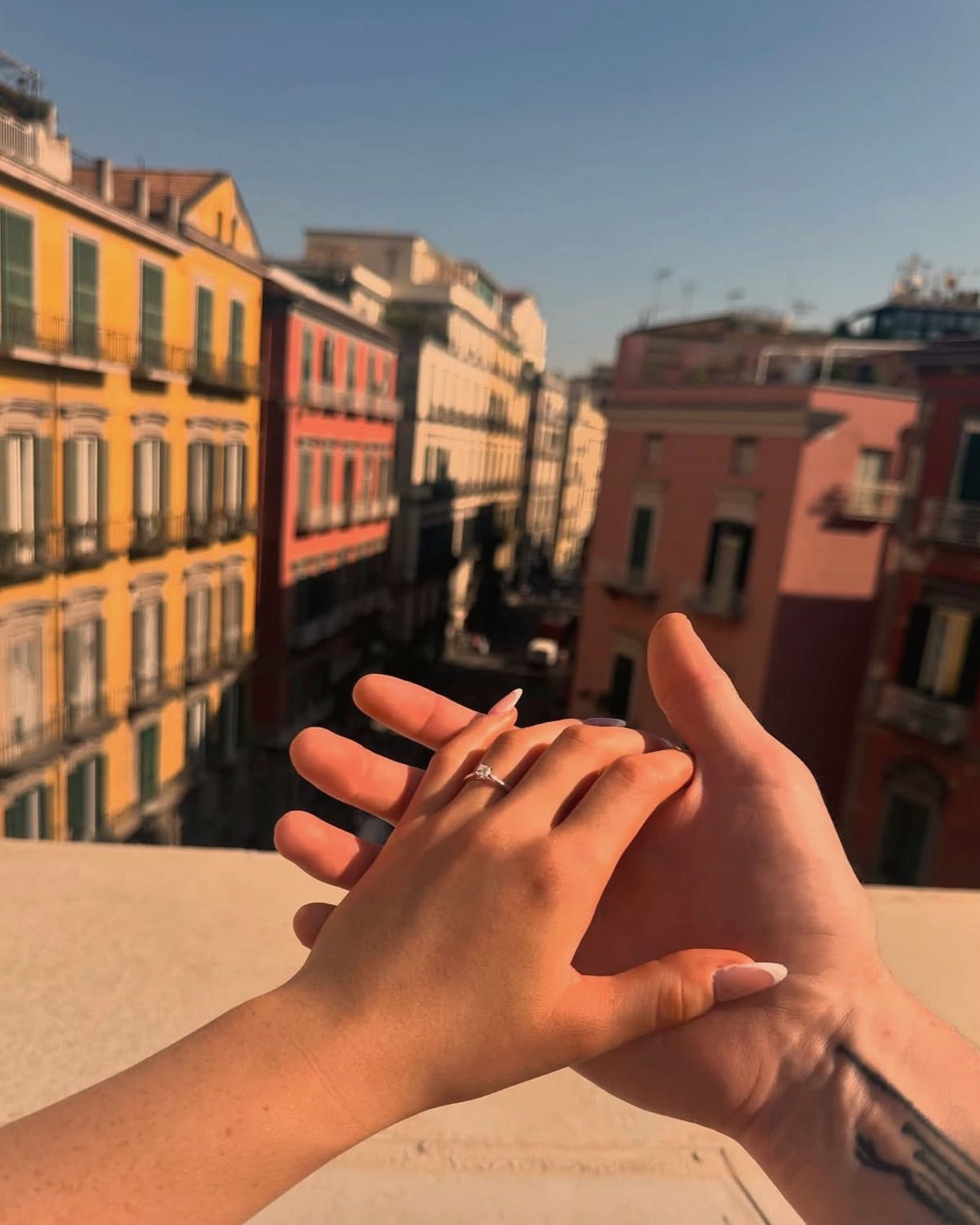 Their hands together on a sunny balcony, the engagement ring on her finger